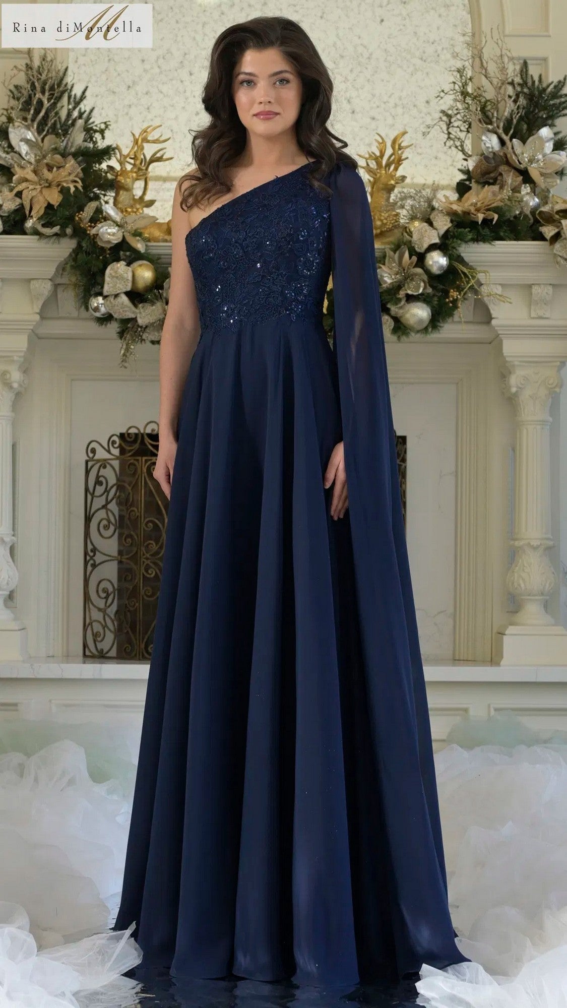 A woman stands indoors in the navy Long Formal Dress: Rina Di Montella RD3354, featuring a one-shoulder design with a floral-embroidered bodice and flowing chiffon A-line skirt; festive decorations appear in the background.